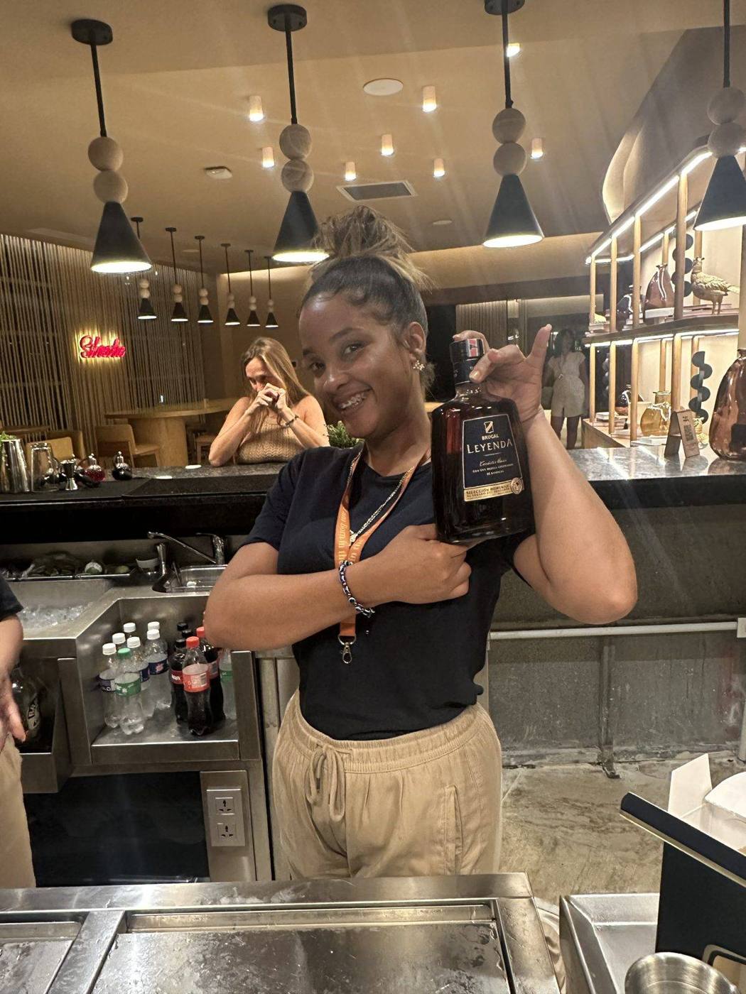 A smiling bartender giving a thumbs up while holding a bottle of Dominican rum behind the bar