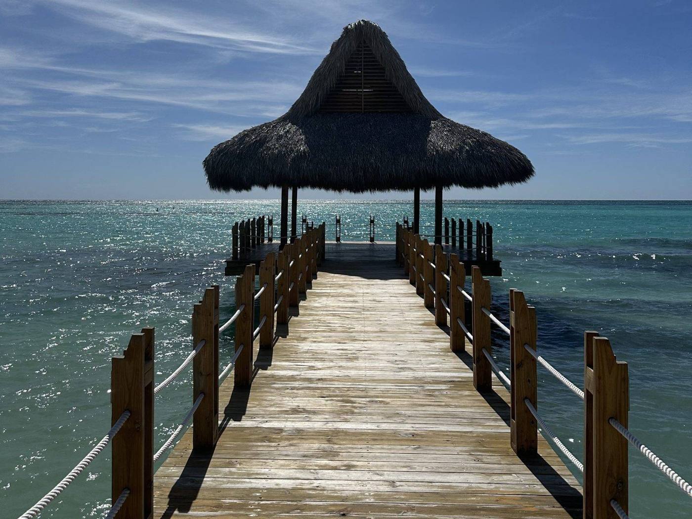 A wooden pier leading out to a thatched gazebo over clear turquoise water under a bright sky
