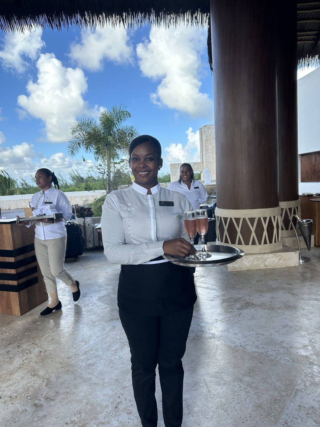 A smiling resort server in a white jacket carrying a tray of drinks in an open-air lobby