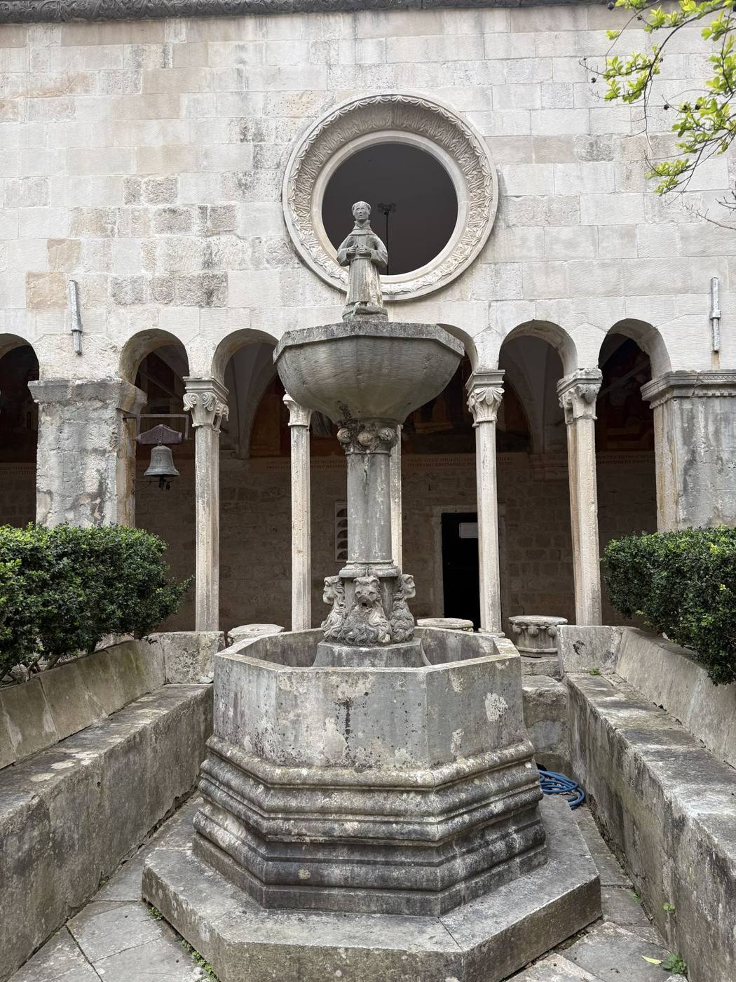 A carved stone fountain in the center of the Franciscan Monastery cloister, surrounded by arched columns and hedges