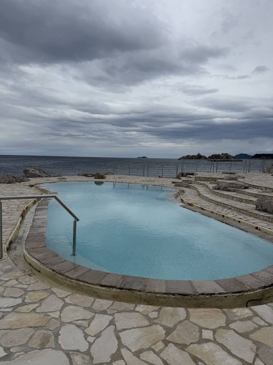 An oval pool set into the stone shoreline beside the sea under cloudy skies