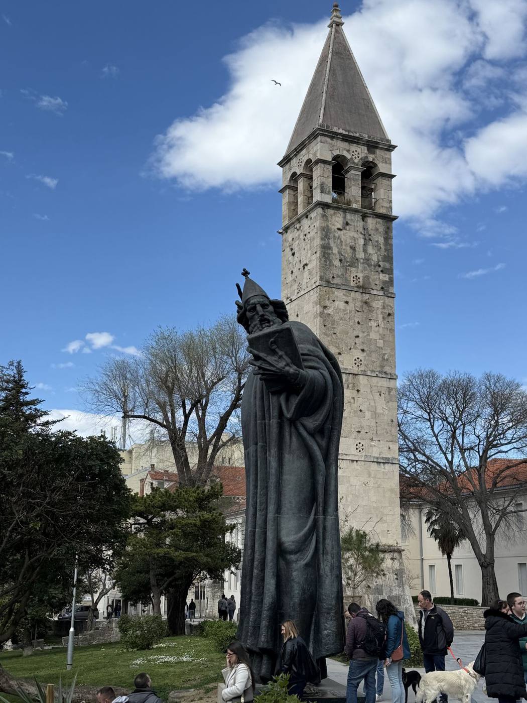 The giant bronze statue of Gregory of Nin beside a stone bell tower and bare trees in Split