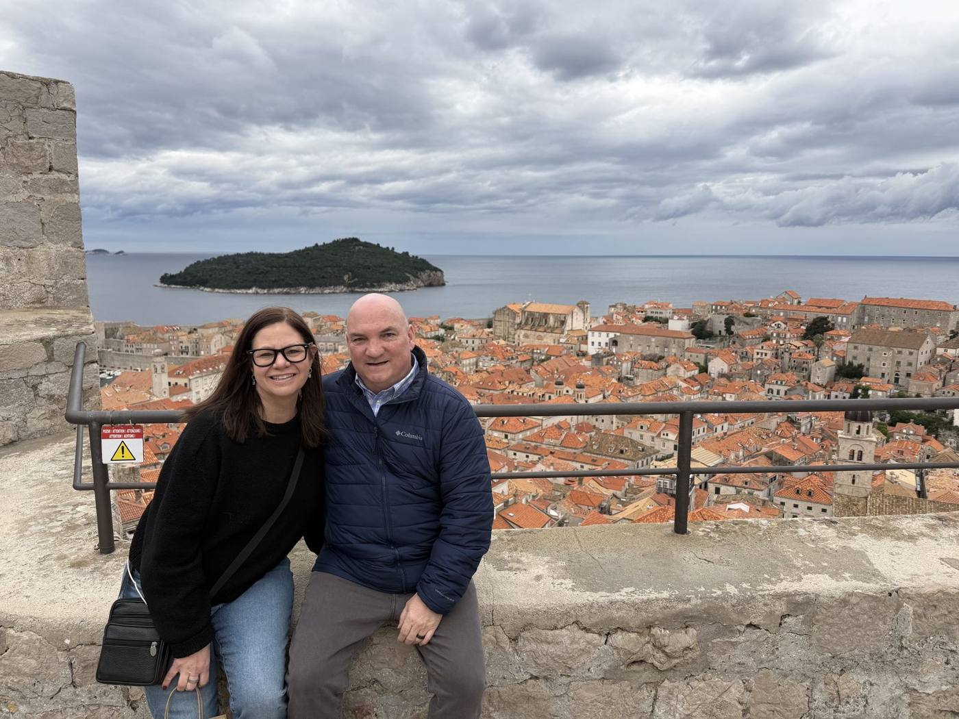 Troy and Mikki sitting on the stone city walls of Dubrovnik, with red rooftops and Lokrum Island behind them