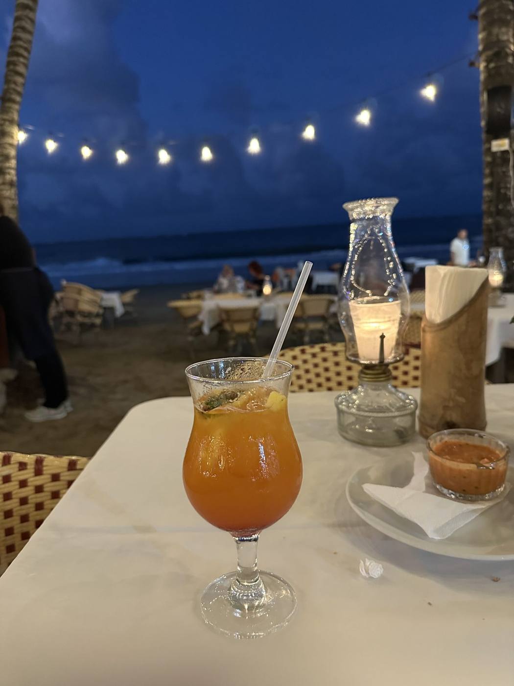 A candlelit dinner table on the sand under string lights with a tropical drink