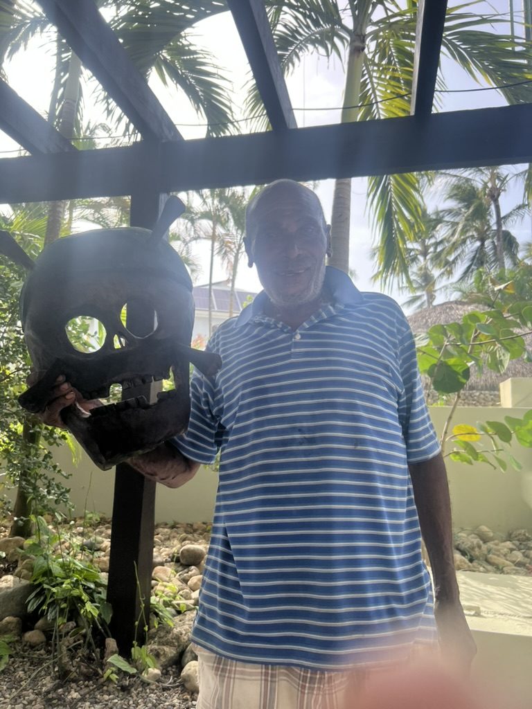 A smiling Dominican woodcarver in a striped polo holding up the large wooden skull he carved
