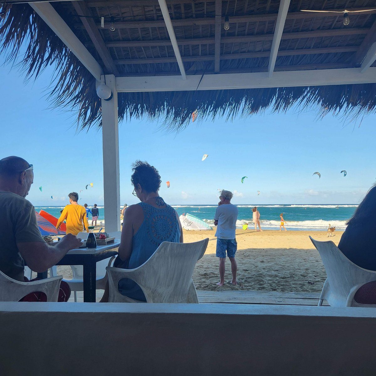 Diners under a thatched roof at a beachfront restaurant, looking out at the sand, the ocean and kite surfers on Kite Beach