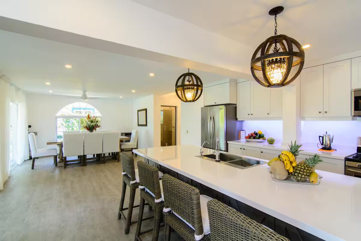 The bright open kitchen at Olas de Oro Villa 25 in Cabarete, with a long white island, wicker bar stools and a dining table for the whole group