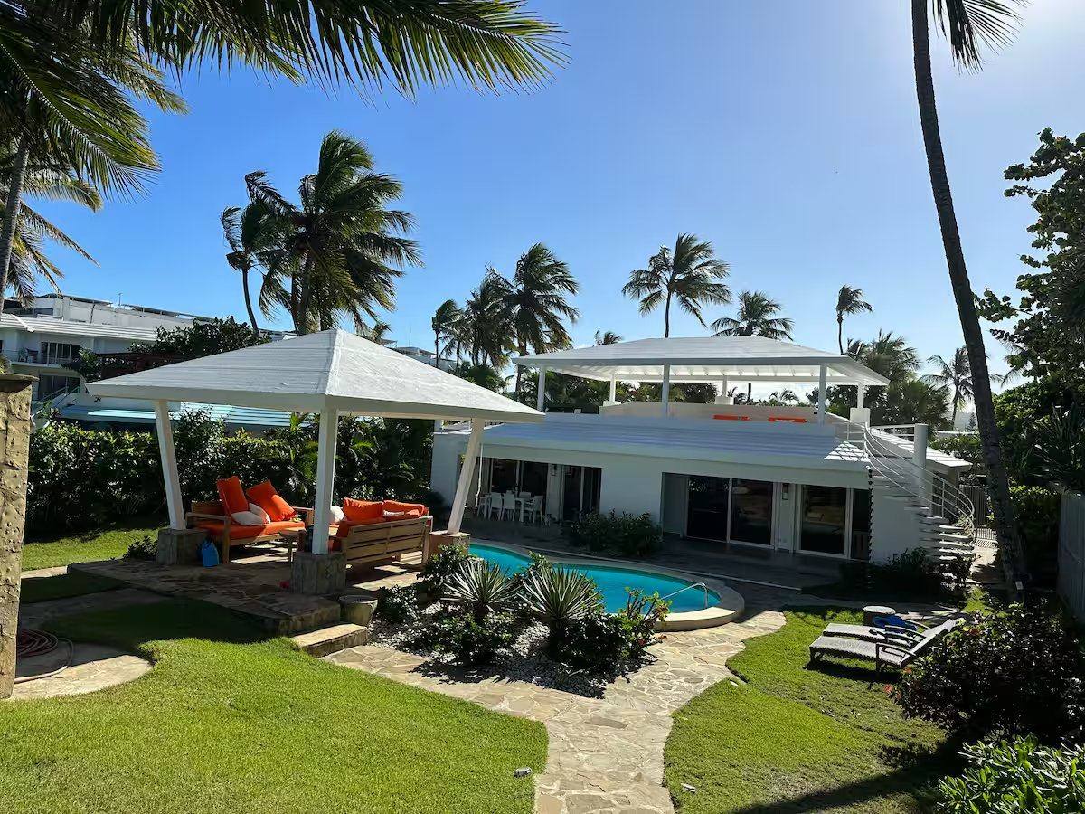 A white beachfront villa with a kidney-shaped pool, a garden cabana with orange cushions and tall palm trees