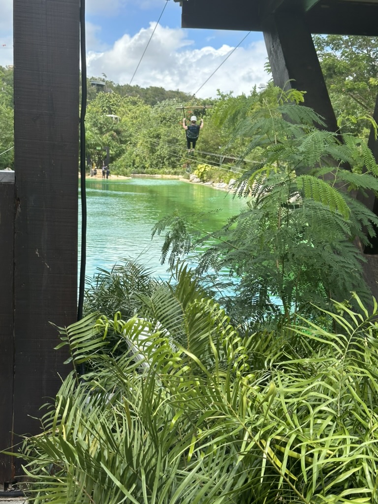 A rider on a zip line flying over a green lagoon surrounded by jungle