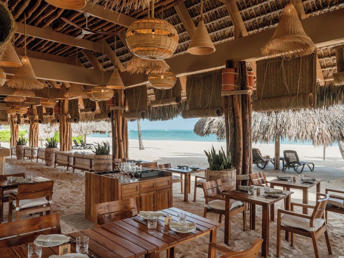 Zel's private beach club restaurant, with wood tables on the sand under a thatched roof and the sea beyond