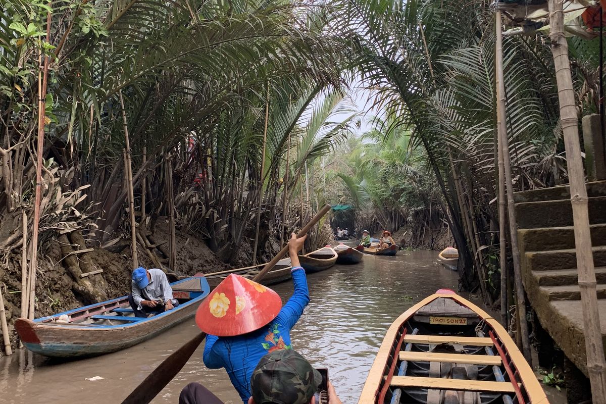 A rower in a conical hat paddles a wooden boat down a palm-lined canal in the Mekong Delta